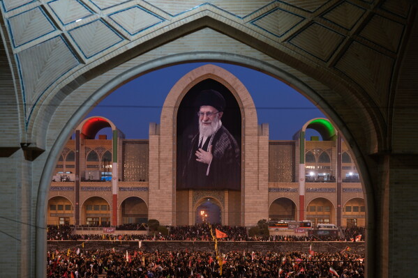 Mourners gather beneath a portrait of the late Iranian Supreme Leader Ayatollah Ali Khamenei during the funeral ceremonies for Khamenei and members of his family at the Imam Khomeini Mosalla Grand Mosque in Tehran, Iran, Saturday, July 4, 2026. (AP Photo/Altaf Qadri)